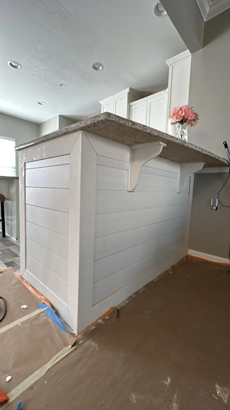 Kitchen peninsula before painting, bare white shiplap cladding and corbels under a granite bar top, floor papered off