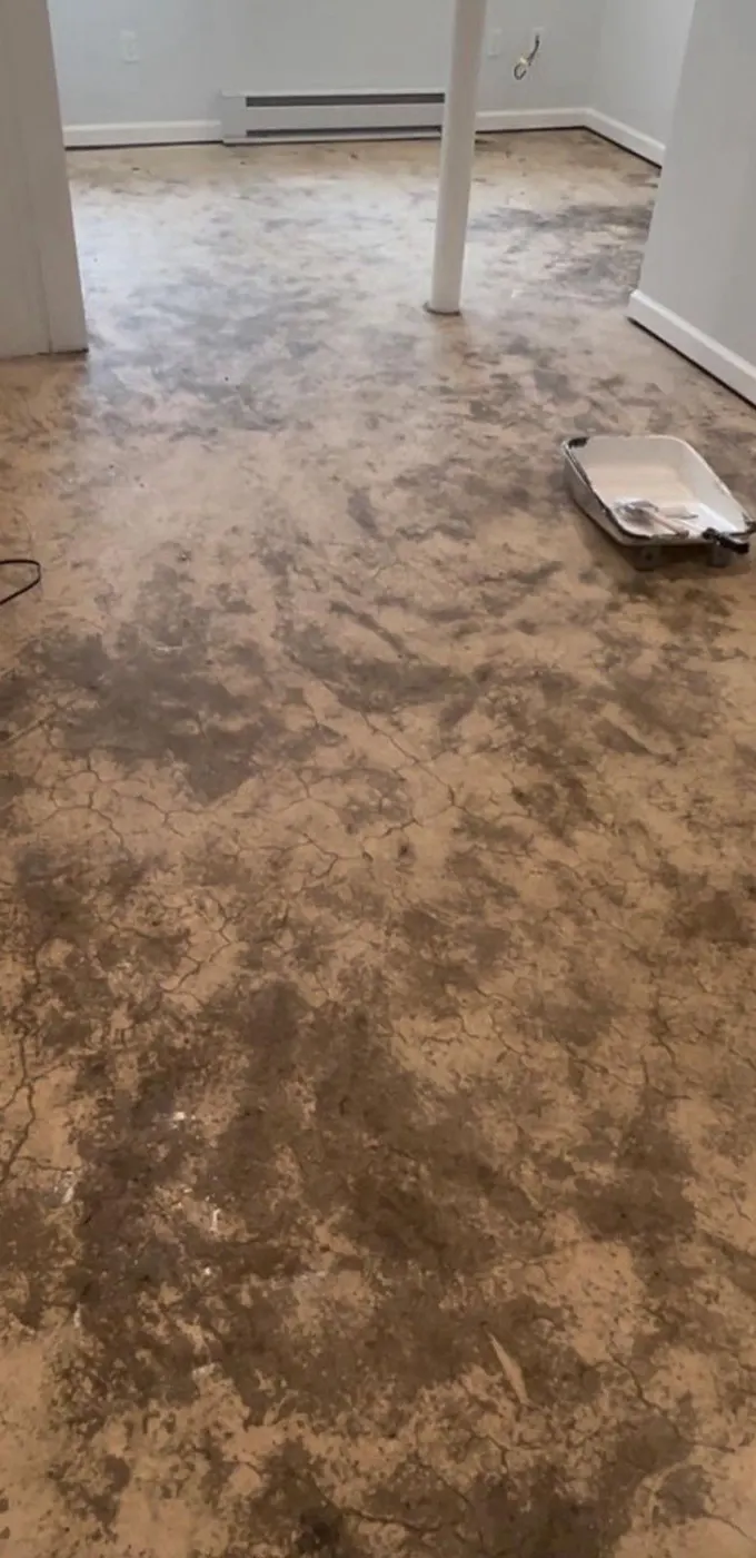 Basement concrete floor before coating, stained and blotchy with a paint tray on the floor and white baseboard heaters along the wall
