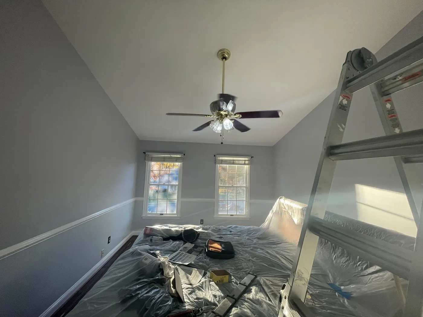 Bedroom mid-job with the furniture under plastic, a stepladder open by the wall and the ceiling fan still up