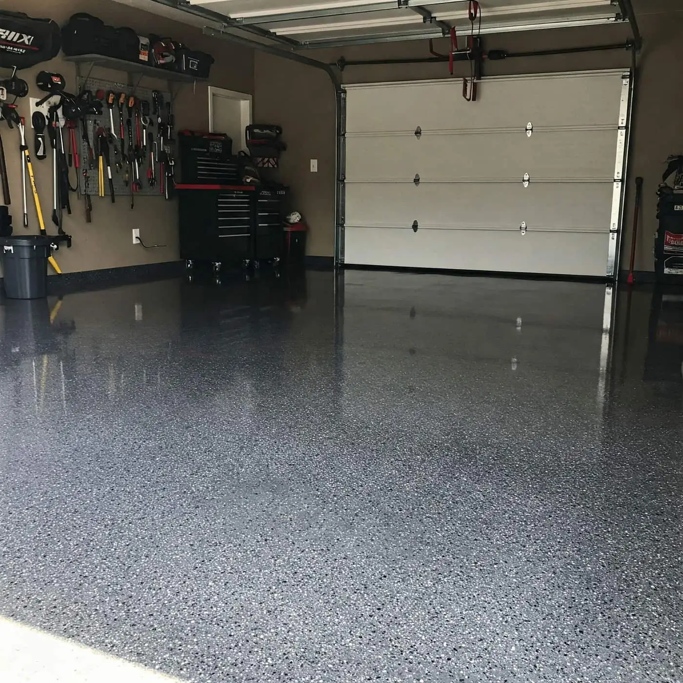 Garage floor finished in a gray and black flake coating, a tool wall and a white roll-up door