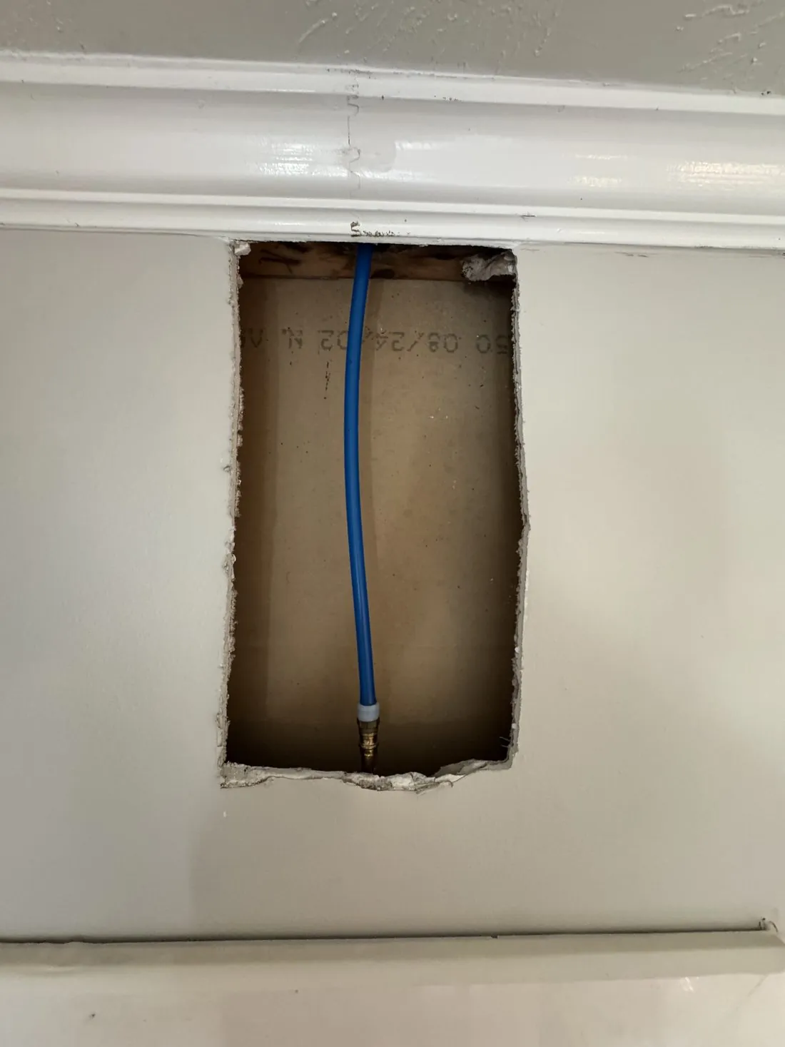Rectangular hole cut in a painted interior wall below the crown molding, exposing the stud bay and a blue water line inside