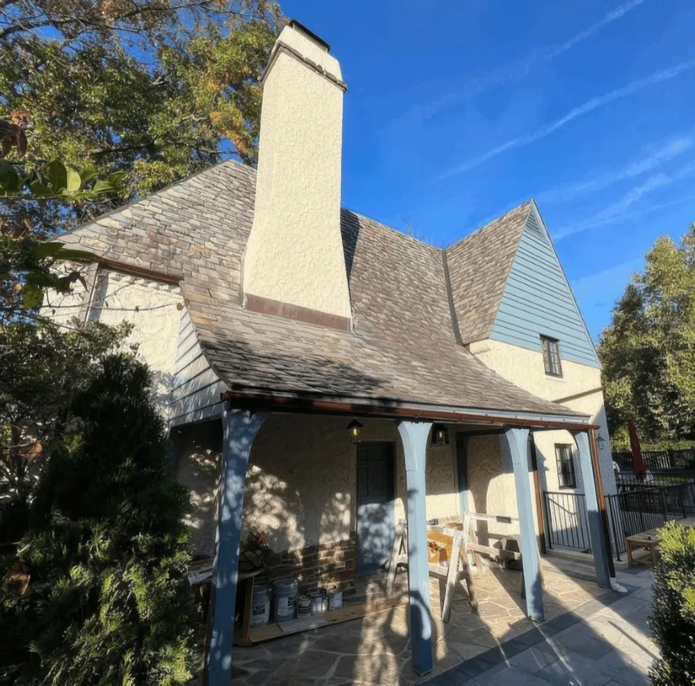 Painted stucco chimney and blue-gray porch posts on a stucco home, paint cans and a sawhorse staged on the stone patio under the porch, by Rise and Prime Painting