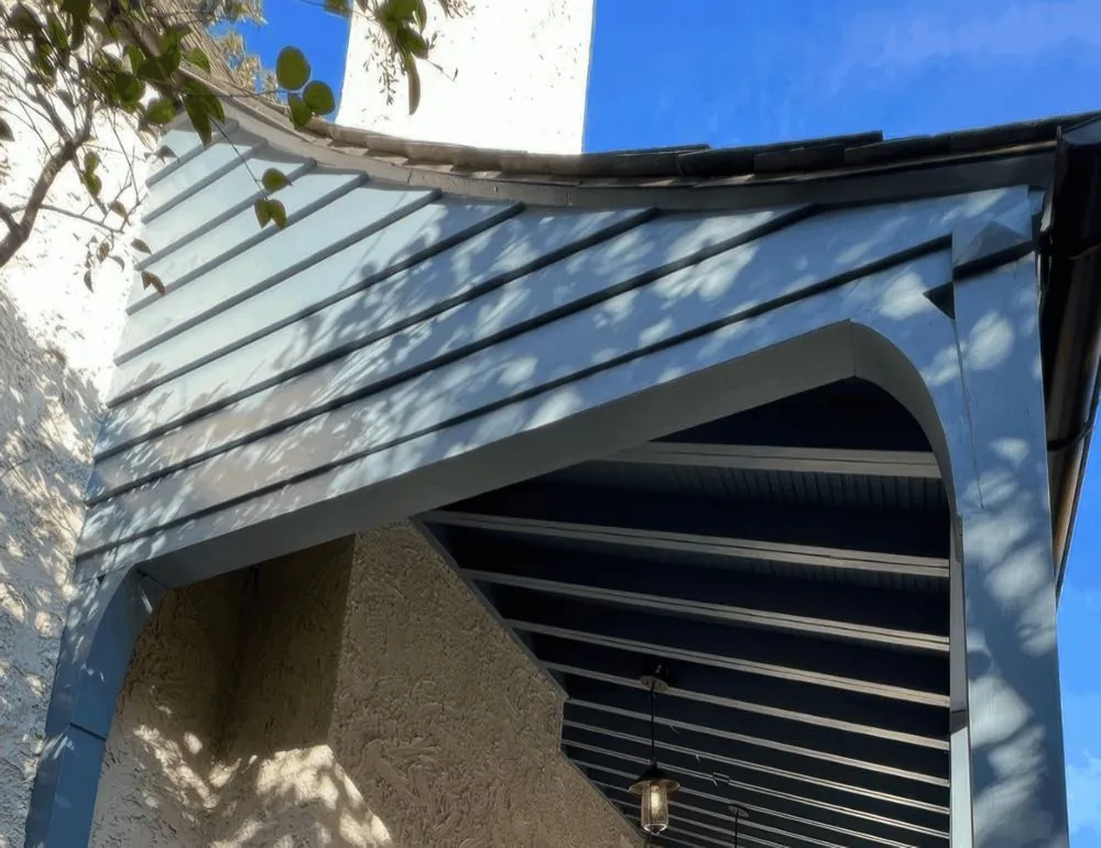 Blue-gray painted beams and arch of a covered entry with a standing-seam metal roof, set against white stucco, photographed from below, by Rise and Prime Painting