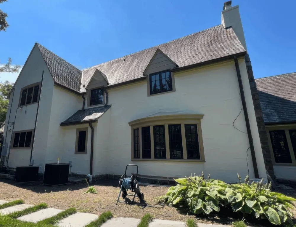 Side and rear elevation of a stucco home painted cream with dark window trim by Rise and Prime Painting, a garden hose reel standing on the path