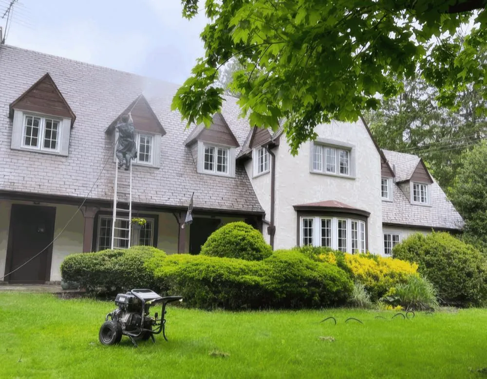Extension ladder set against the upper elevation of a large painted-brick home with dormers, pressure washer staged on the lawn, mid-project for Rise and Prime Painting
