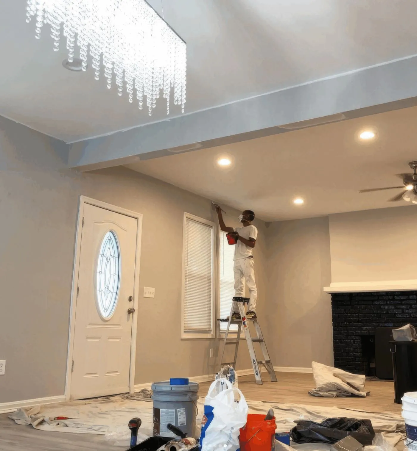 Rise and Prime Painting crew member on a step ladder cutting in the wall-to-ceiling line of a living room by hand, drop cloths down and buckets staged on the floor