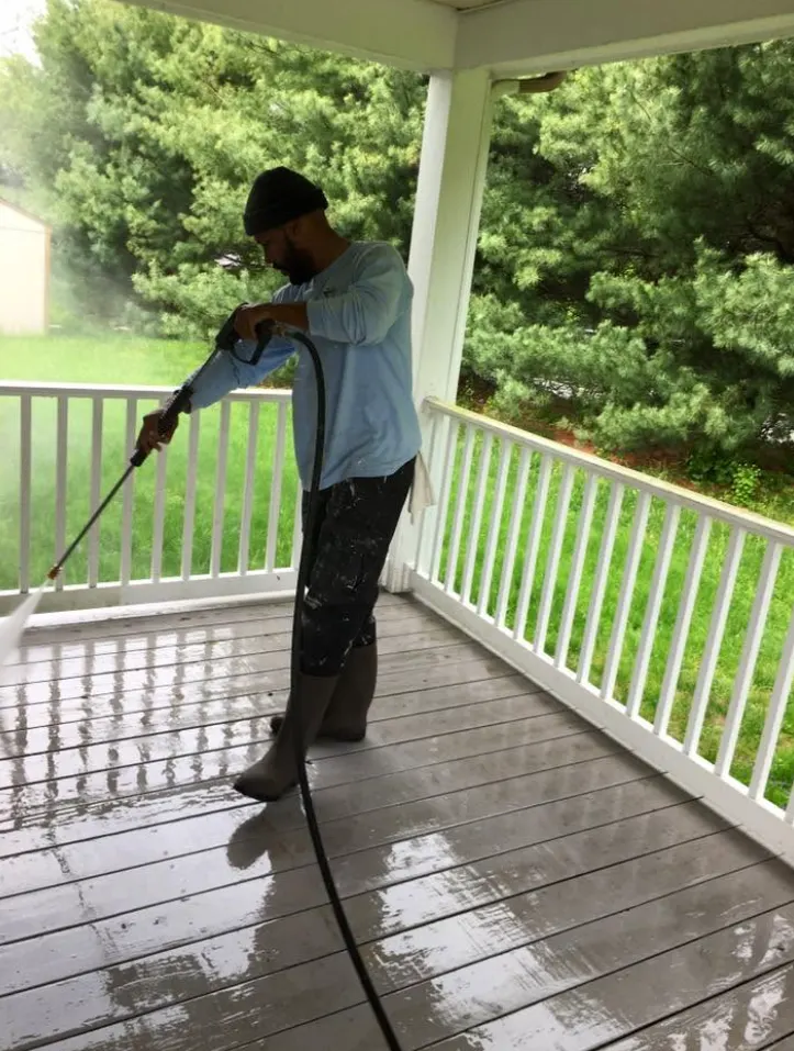 Ray Gomez of Rise and Prime pressure washing a wet gray painted porch floor with a hose and wand, white painted railings along both sides and green pine trees and lawn beyond