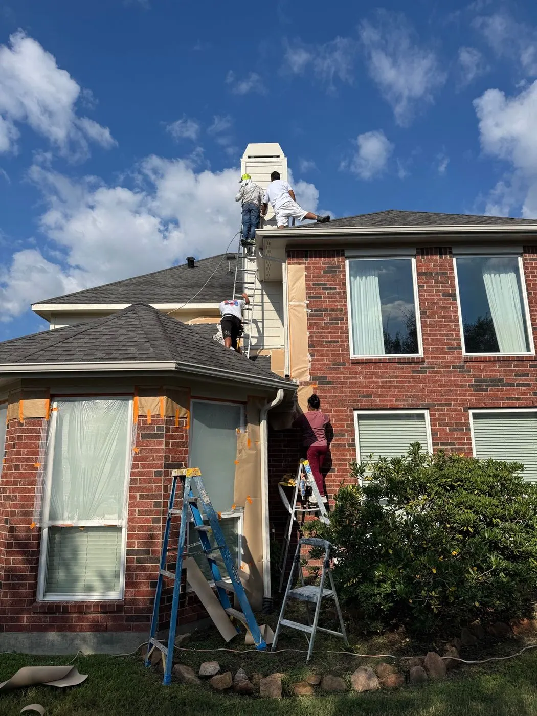 Painters working at three heights on one brick house: two on the roof at the upper siding, one on a ladder at the bay window and one at the gutter line