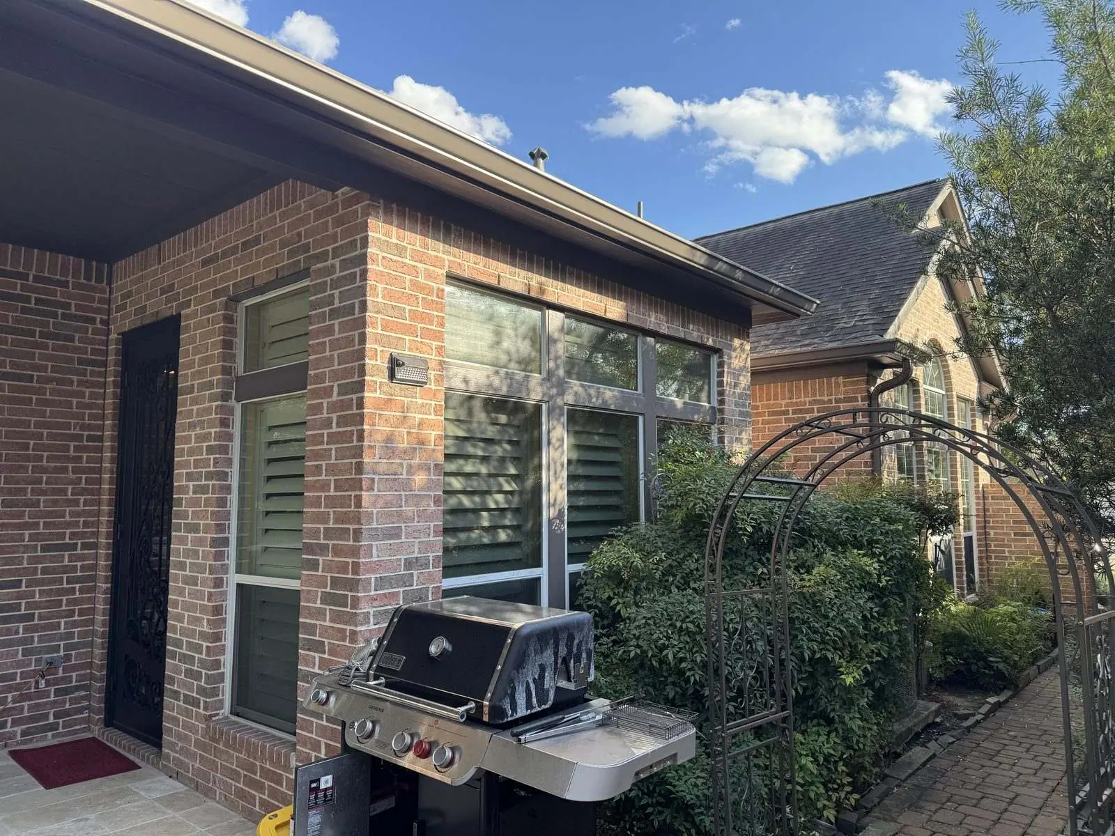 Side elevation of a red brick home with repainted window trim and a dark brown eave, a garden arch and grill in the foreground