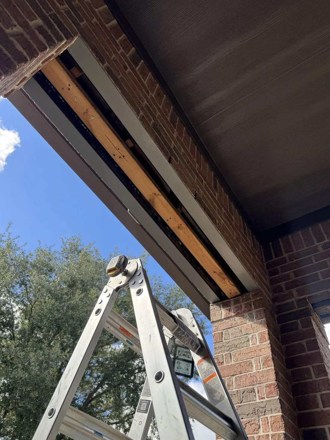 Extension ladder set against the eave of a brick home where the fascia has been taken off, the exposed framing visible against the finished dark soffit
